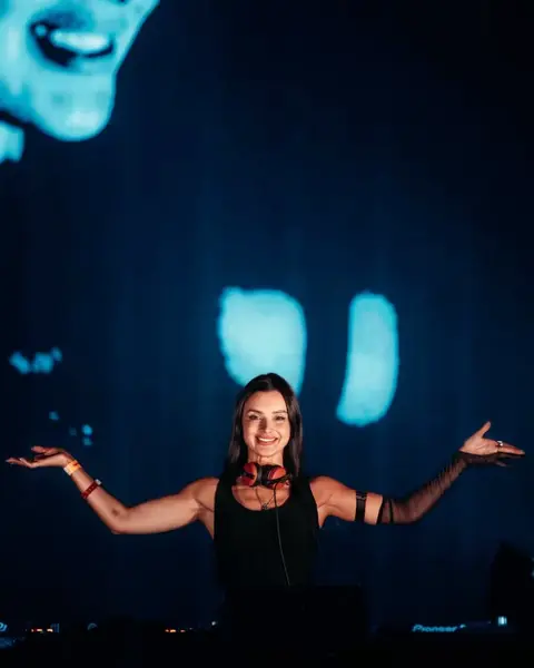 Korolova smiling, headphones around her neck and arms outstretched, in front of a blue LED screen.